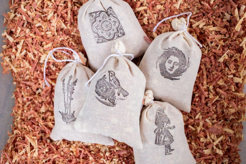 May include: Five off-white fabric drawstring pouches featuring black ink illustrations. The designs include a geometric tile pattern, a bird in a nest, a sprig of lavender, a human face surrounded by swirling lines, and a standing figure in a dress.
