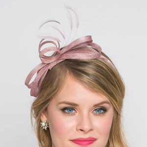 May include: A dusty rose-colored headband with a bow and feather accents. The headband is made of a textured fabric and is designed to be worn on the head. The accessory is suitable for special occasions.