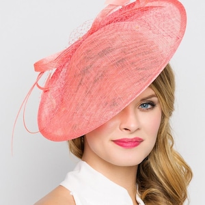 May include: A coral-colored hat with a wide brim and netting detail. The hat features a decorative bow and delicate feather accents. This elegant headwear is suitable for special occasions and events.