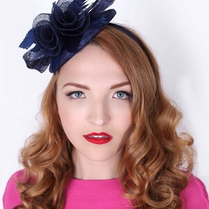 K&ouml;nnte beinhalten: Ein marineblauer Fascinator mit Blumenmuster und Federakzent. Der Fascinator wird auf dem Kopf einer Frau getragen.