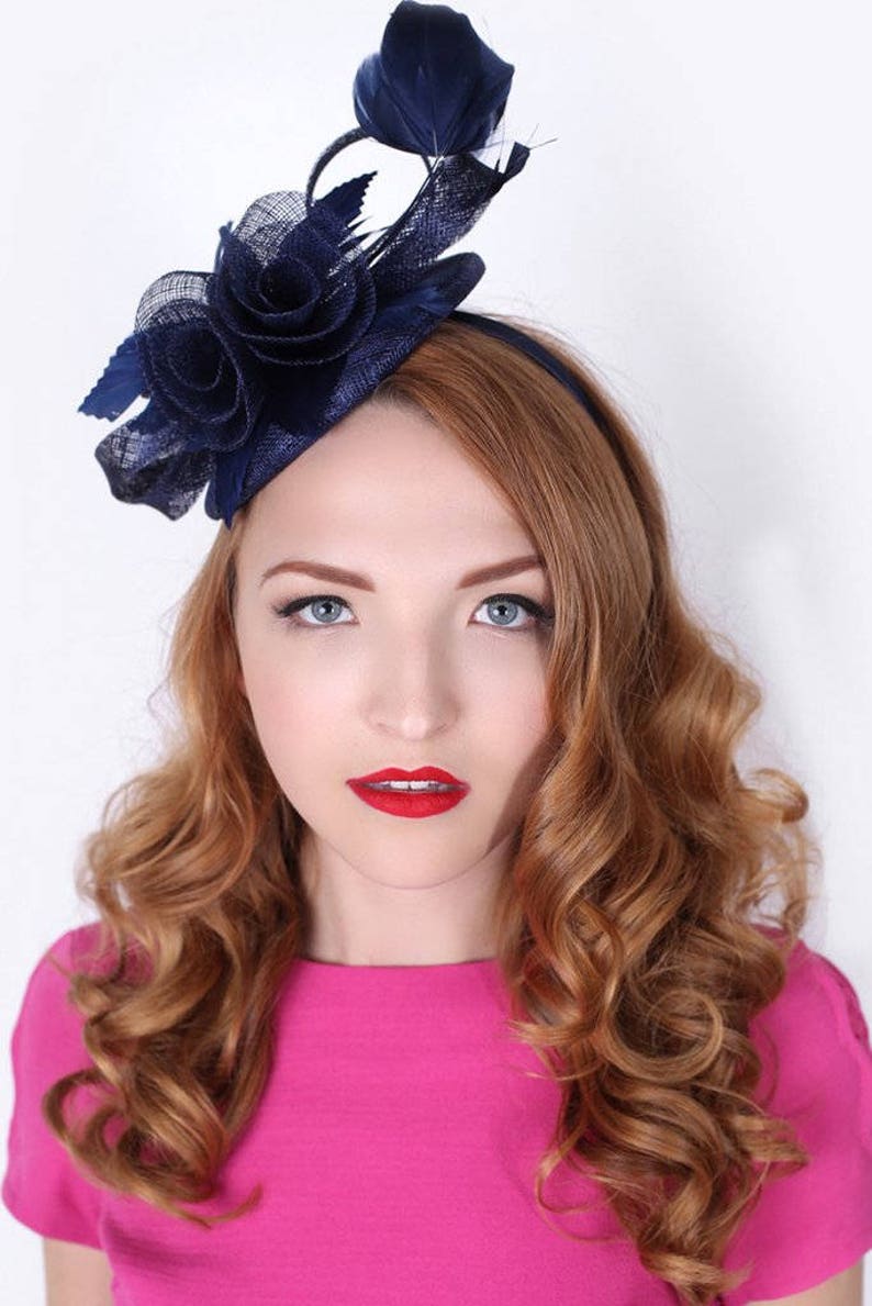 K&ouml;nnte beinhalten: Ein marineblauer Fascinator mit Blumenmuster und Federakzent. Der Fascinator wird auf dem Kopf einer Frau getragen.