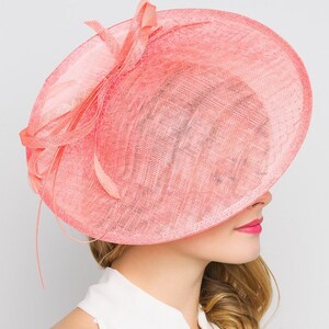May include: A coral-colored, wide-brimmed hat with a delicate, woven texture. The hat features a decorative bow and feather accents. The brim is large and covers the face, creating a stylish and elegant look. Perfect for special occasions.