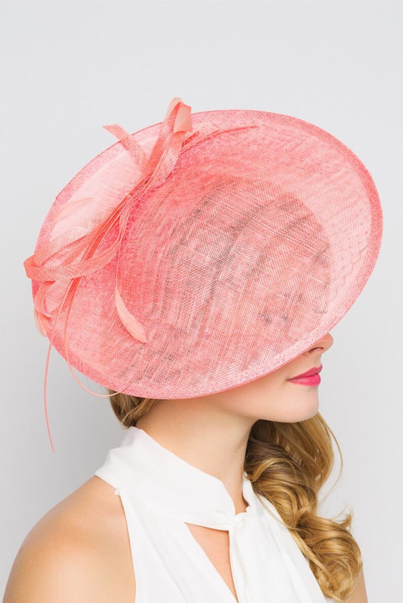 May include: A coral-colored, wide-brimmed hat with a delicate, woven texture. The hat features a decorative bow and feather accents. The brim is large and covers the face, creating a stylish and elegant look. Perfect for special occasions.