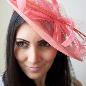 May include: A pink hat with a wide brim and decorative ribbons, netting, and feathers. The hat is a light coral color and is worn on the head. The hat is a formal accessory, suitable for events like weddings or the Kentucky Derby.