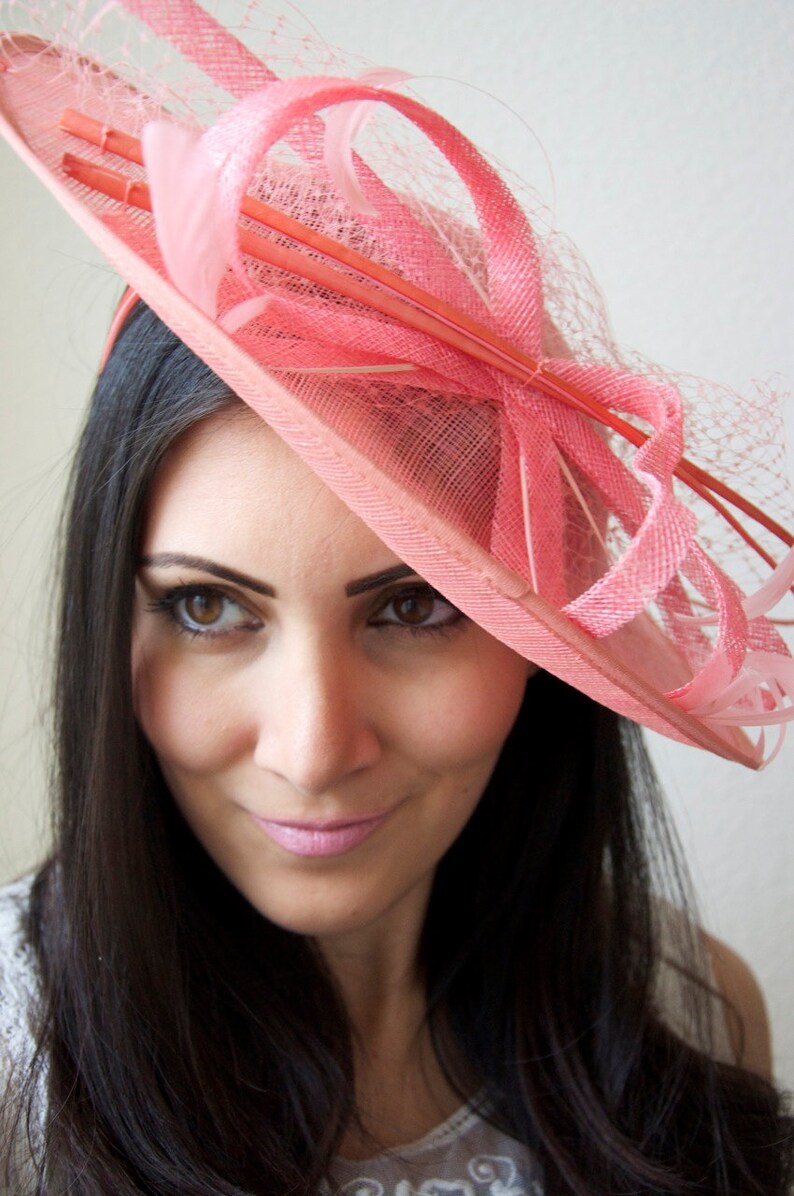 May include: A pink hat with a wide brim and decorative ribbons, netting, and feathers. The hat is a light coral color and is worn on the head. The hat is a formal accessory, suitable for events like weddings or the Kentucky Derby.