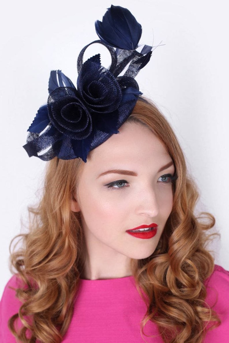 K&ouml;nnte beinhalten: Ein marineblauer Fascinator mit einer gro&szlig;en Blume und Federn. Der Fascinator wird auf dem Kopf einer Frau getragen.