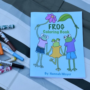 May include: A light blue "FROG Coloring Book" with a cartoon frog illustration and the text "By: Hannah Moyer". Assorted colorful markers are scattered on a striped surface.