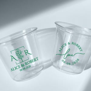 May include: Two clear plastic cups with green text. One cup features the initials "A R" inside a square with floral accents, and the names "Alice & Robert" with the date "06.08.2026". The other cup has the word "love" and the same names and date.