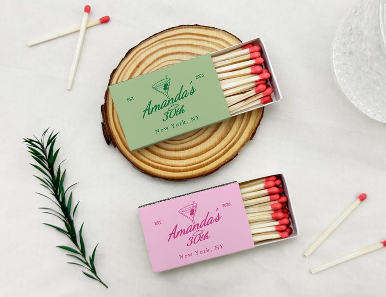 May include: Two matchboxes, one green and one pink, with the text "Amanda's 30th New York, NY" and a martini glass logo. The matchboxes are filled with wooden matches with red tips. The boxes rest on a wooden slice.