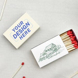May include: Two rectangular matchboxes with wooden matches featuring red tips. One box is cream-colored with blue text reading YOUR DESIGN HERE. The other box is white with a black line drawing of a house and is partially open, revealing the matches.