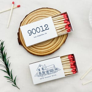 May include: Two white matchboxes with red-tipped matches are displayed on a wooden slice. One box has the text "90012 Los Angeles, CA" and the other has a house illustration and "5678 Sunset Blvd." Scattered matches and greenery complete the scene.