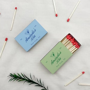 May include: Two matchboxes, one light blue and one green, with the text "Amanda's 30th New York, NY" and a cocktail glass illustration. Several matches with red tips are scattered around the boxes.