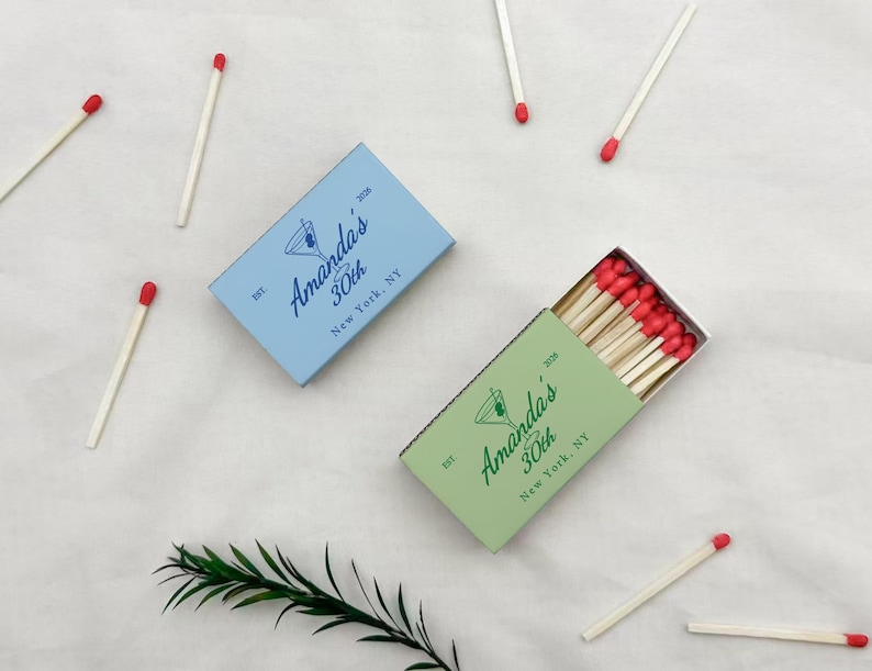 May include: Two matchboxes, one light blue and one green, with the text "Amanda's 30th New York, NY" and a cocktail glass illustration. Several matches with red tips are scattered around the boxes.