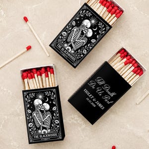 May include: Two black matchboxes with red-tipped wooden matches. One box features a white illustration of two skeletons kissing surrounded by flowers, the sun, and moon, with the text THE BLACKWOODS. The other box displays the text TILL DEATH DO US PART, VIOLET & JAMES, 10.31.2028.