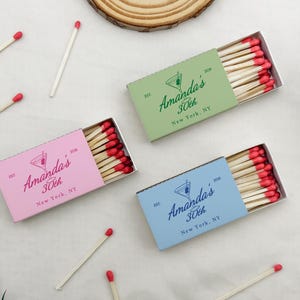 May include: Three matchboxes in pink, green, and blue, each with a custom design for a 30th birthday. The boxes contain wooden matches with red tips. The text on the boxes reads "Amanda's 30th New York, NY."