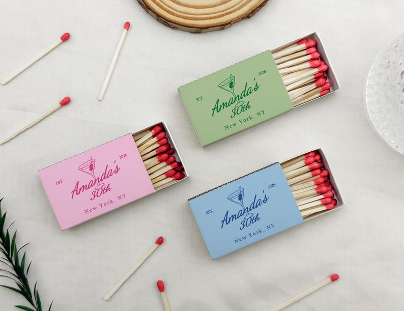 May include: Three matchboxes in pink, green, and blue, each with a custom design for a 30th birthday. The boxes contain wooden matches with red tips. The text on the boxes reads "Amanda's 30th New York, NY."