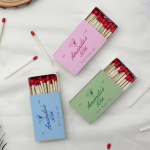 May include: Three matchboxes in pink, green, and blue, each filled with wooden matches with red tips. The boxes are labeled "Amanda's 30th New York, NY" with a cocktail glass illustration. Several loose matches are scattered around.