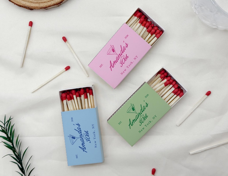 May include: Three matchboxes in pink, green, and blue, each filled with wooden matches with red tips. The boxes are labeled "Amanda's 30th New York, NY" with a cocktail glass illustration. Several loose matches are scattered around.