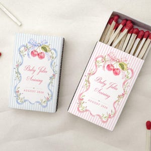 May include: Two rectangular matchboxes featuring a cherry and ribbon design. One box is blue-striped, the other pink-striped. Both display the text Baby Jalia Arriving and AUGUST 2026. The open box reveals wooden matches with red tips, surrounded by loose matches.