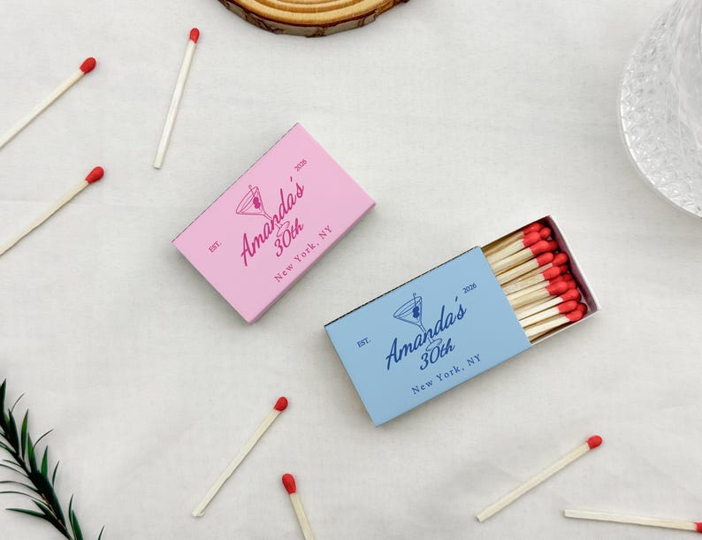 May include: Two matchboxes, one pink and one blue, with the text "Amanda's 30th New York, NY" and a cocktail glass logo. Several matches with red tips are scattered around. The pink box is closed, while the blue box is open, revealing the matches.
