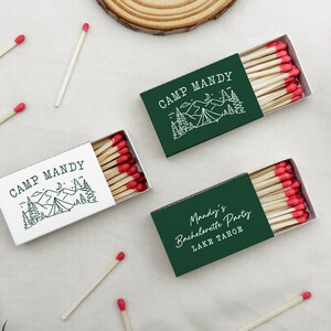 May include: Matches in decorative boxes with red tips. The boxes are white and dark green, with the text "CAMP MANDY" and a mountain and tent design. One box says "Mandy's Bachelorette Party LAKE TAHOE".