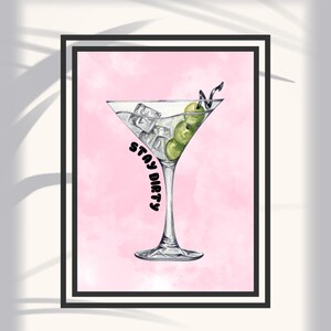 Può includere: Stampa incorniciata con un'illustrazione ad acquerello di un bicchiere da martini pieno di cubetti di ghiaccio e olive verdi. Le parole "STAY DIRTY" sono stampate verticalmente sul bicchiere. Lo sfondo è un morbido lavaggio rosa.