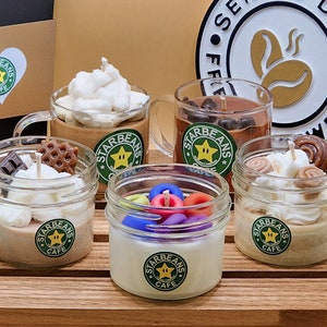 May include: An assortment of six small glass jars with lids, each containing a different scented candle. The candles are decorated with various toppings, including whipped cream, chocolate chips, and sprinkles. The jars are labelled "Starbeans Cafe".