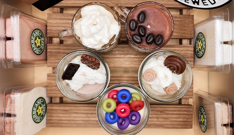 May include: Six glass jars with lids containing different scented wax melts. The wax melts are arranged in a wooden crate. The wax melts are shaped like coffee cups, doughnuts, and other treats. The jars are labelled "Star Beans Cafe".