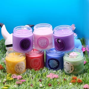 May include: Six small glass candle jars with colorful wax and labels featuring cartoon animals. The candles are arranged in a semi-circle on a bed of green grass.