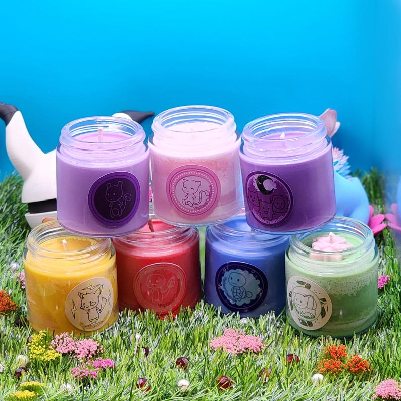May include: Six small glass candle jars with colorful wax and labels featuring cartoon animals. The candles are arranged in a semi-circle on a bed of green grass.