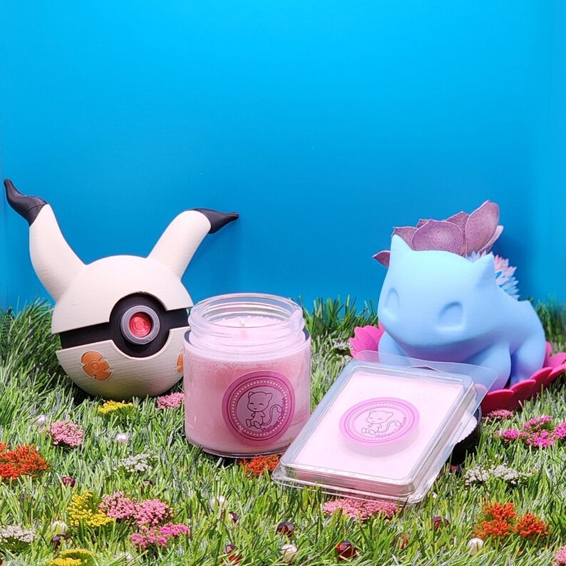 May include: Two pink candles with a white cat logo on the front, one in a glass jar and one in a clear plastic container, sitting on a bed of green grass with pink flowers. A blue ceramic figurine of a cat is sitting on a pink flower pot behind the candles.