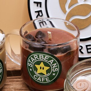 May include: A brown coffee-scented candle in a clear glass mug with a handle. The candle has a white wick and is decorated with chocolate coffee beans. The mug has a green and white sticker that says "Starbeans Cafe" with a yellow star and the text "Vigilant Creations".