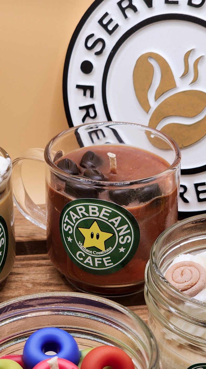 May include: A brown coffee-scented candle in a clear glass mug with a handle. The candle has a white wick and is decorated with chocolate coffee beans. The mug has a green and white sticker that says "Starbeans Cafe" with a yellow star and the text "Vigilant Creations".