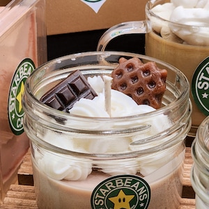 May include: A glass jar candle with a brown, white and tan wax base. The candle is topped with white whipped cream, a chocolate bar and a waffle cookie. The jar has a green and yellow sticker with the text "Starbeans Cafe" and a star with a face.