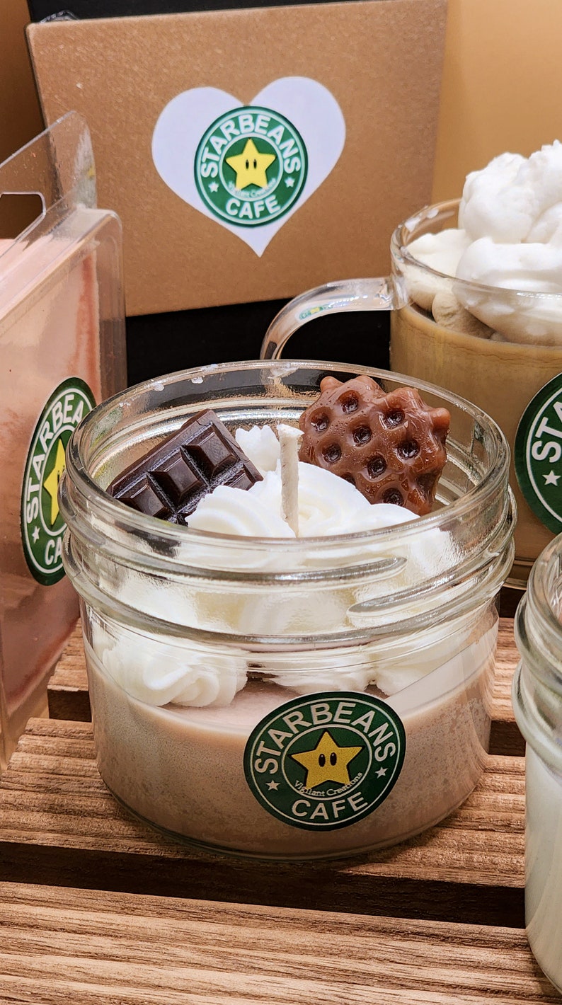 May include: A glass jar candle with a brown, white and tan wax base. The candle is topped with white whipped cream, a chocolate bar and a waffle cookie. The jar has a green and yellow sticker with the text "Starbeans Cafe" and a star with a face.