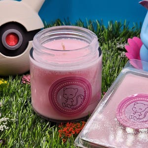 May include: A pink candle in a glass jar with a pink label featuring a cartoon cat. The label says "Mewtwo Candles". The candle is sitting on a bed of green artificial grass.
