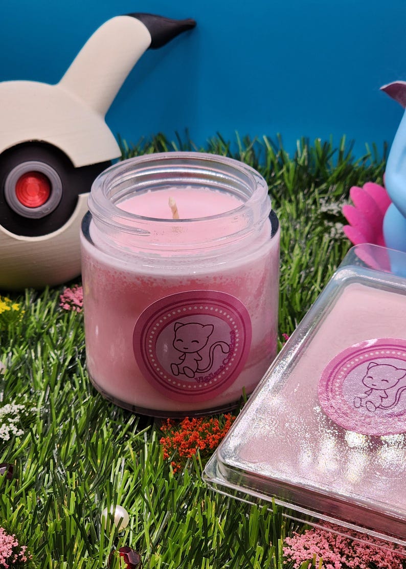 May include: A pink candle in a glass jar with a pink label featuring a cartoon cat. The label says "Mewtwo Candles". The candle is sitting on a bed of green artificial grass.