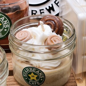 May include: A close-up of a candle in a glass jar with a white frosting-like topping and brown swirl sweets. The jar has a green label with the text "Starbeans Cafe" and a yellow star with a green outline.