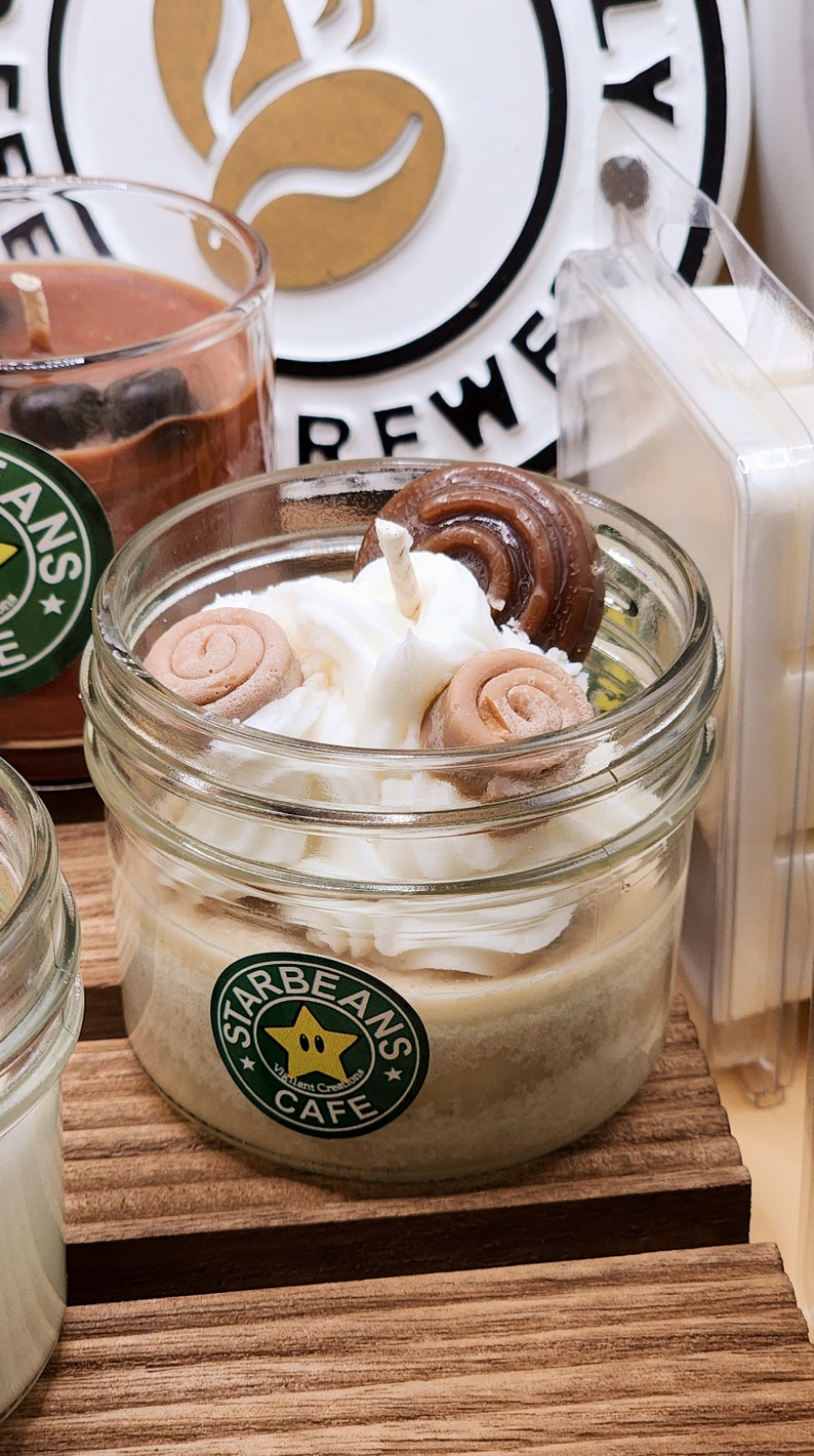 May include: A close-up of a candle in a glass jar with a white frosting-like topping and brown swirl sweets. The jar has a green label with the text "Starbeans Cafe" and a yellow star with a green outline.