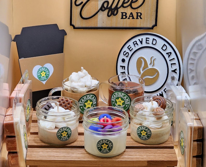 May include: An assortment of six small glass jars with lids, each containing a different scented candle. The candles are decorated with various toppings, including whipped cream, chocolate chips, and sprinkles. The jars are labelled "Starbeans Cafe".