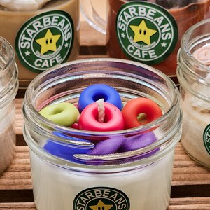 May include: Four glass jars containing candles. The candles are topped with colourful donut-shaped wax. The jars have green and white labels with the text "Starbeans Cafe" and a yellow star with a face.