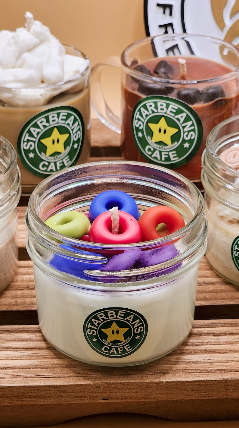 May include: Four glass jars containing candles. The candles are topped with colourful donut-shaped wax. The jars have green and white labels with the text "Starbeans Cafe" and a yellow star with a face.