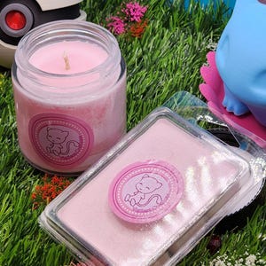 May include: Pink scented candle in a glass jar with a pink and white label featuring a cartoon pink cat. The candle is sitting on a bed of green grass with a pink wax melt in a clear plastic container next to it.
