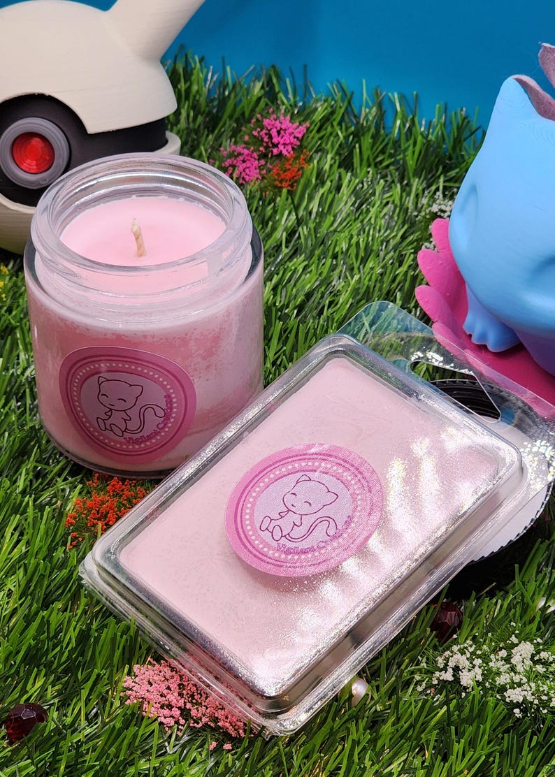 May include: Pink scented candle in a glass jar with a pink and white label featuring a cartoon pink cat. The candle is sitting on a bed of green grass with a pink wax melt in a clear plastic container next to it.
