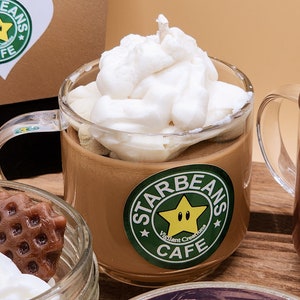 May include: A glass mug filled with a brown liquid topped with whipped cream and a white wick. The mug has a green and yellow sticker that says "Starbeans Cafe" with a yellow star with a face.