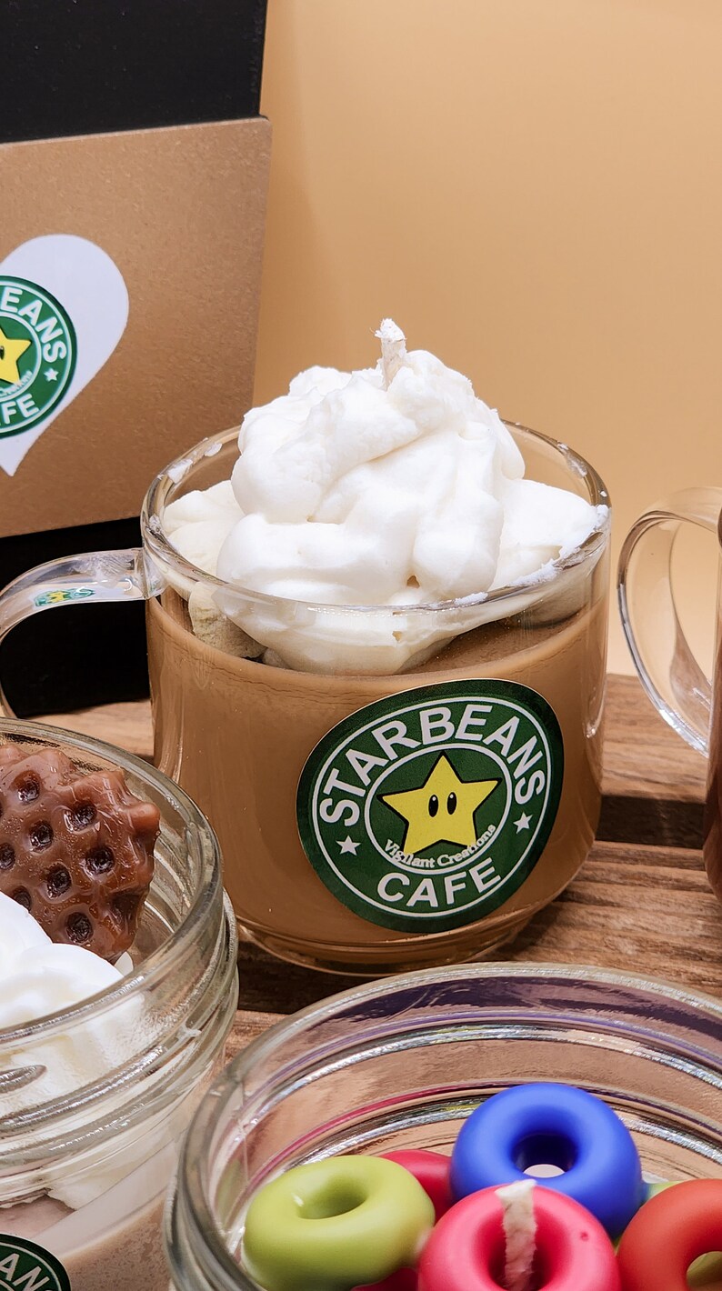 May include: A glass mug filled with a brown liquid topped with whipped cream and a white wick. The mug has a green and yellow sticker that says "Starbeans Cafe" with a yellow star with a face.