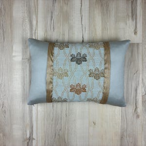 May include: A rectangular decorative pillow with a light blue background and a central patterned panel. The panel features a light blue background with a floral design in gold and brown. Gold trim borders the patterned panel.