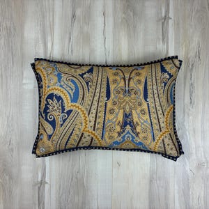 May include: Rectangular cushion featuring a paisley design in blue, gold, and brown hues. It has a dark blue border with a dotted trim. The cushion is placed on a light wood-grain surface.