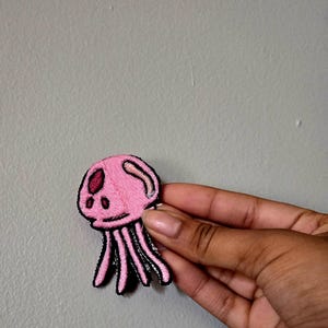 May include: Pink jellyfish patch with black outline and details. The embroidered patch features a rounded head with two eye spots and trailing tentacles. The patch is likely for clothing or accessories.