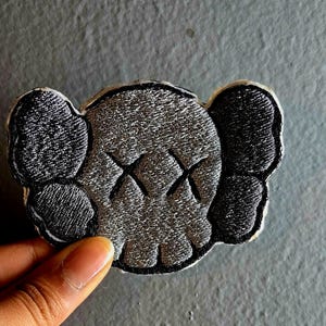 May include: Embroidered patch featuring a stylized skull design with crossed-out eyes. The patch has a gray and black color scheme with a white border. The design includes large ear-like shapes on either side of the skull.